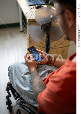Disabled guy in wheelchair checking weather forecast in mobile app on smartphone at home. Heat wave Disabled guy in wheelchair checking weather forecast in mobile app on smartphone at home. Heat wave 117160399