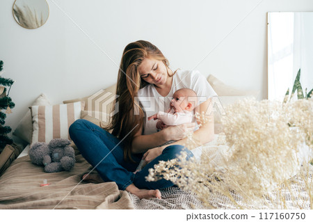 A tender caring mother nurses a newborn baby in her arms while sitting on the bed.	 117160750