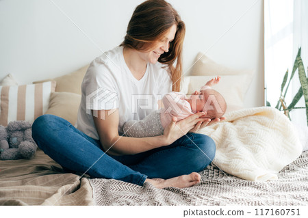 A tender caring mother nurses a newborn baby in her arms while sitting on the bed. A tender caring mother nurses a newborn baby in her arms while sitting on the bed. 117160751