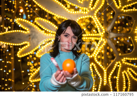 Russian 48 years old woman in blue sweater holding tangerines against yellow christmas lights before Christmas Russian 48 years old woman in blue sweater holding tangerines against yellow christmas lights before Christmas 117160795