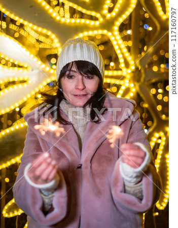 Russian 48 years old woman in blue sweater and pink coat standing against yellow christmas lights before Christmas and holding lit sparklers Russian 48 years old woman in blue sweater and pink coat standing against yellow christmas lights before Christmas and holding lit sparklers 117160797