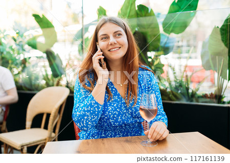 Beautiful woman with glass of wine talking by mobile phone outdoors. Female Waiting for food order, beverage, food and drink concept, happy lunch time banner, enjoy life in public place 117161139