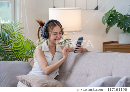 Young Woman Relaxing on Sofa with Headphones and Smartphone in Modern Living Room with Green Plants 117161196