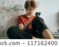 A teenage girl sitting on a bed in a room uses a mobile phone for online communication.	 117162400
