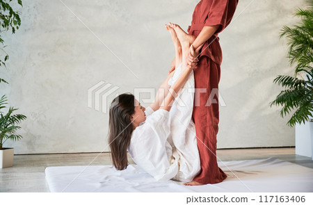 Masseuse making thai yoga massage. Cropped view of woman therapist doing traditional massage treatment, stretching female patient's body. Client lying on mat, enjoying hand massage in calm atmosphere. Masseuse making thai yoga massage. Cropped view of woman therapist doing traditional massage treatment, stretching female patient's body. Client lying on mat, enjoying hand massage in calm atmosphere. 117163406