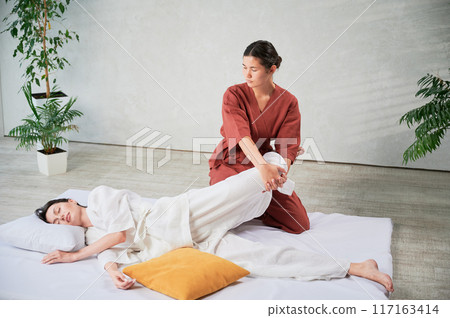 Masseuse making thai yoga massage. Woman therapist doing traditional massage treatment, stretching female patient's body. Client lying on mat, enjoying hand massage in calm atmosphere. 117163414
