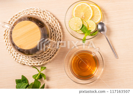 Top view of glass teapot with wooden lid, cup of tea, lemon and lime slices and fresh mint on wooden table Top view of glass teapot with wooden lid, cup of tea, lemon and lime slices and fresh mint on wooden table 117163507