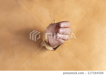 Hand coming out of a hole in a brown paper with copy space. Closeup Hand coming out of a hole in a brown paper with copy space. Closeup 117164500
