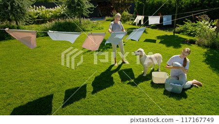 A woman and a girl hang laundry on a clothesline in a sunlit backyard, accompanied by a Golden Retriever, creating a peaceful and homey scene. A woman and a girl hang laundry on a clothesline in a sunlit backyard, accompanied by a Golden Retriever, creating a peaceful and homey scene. 117167749