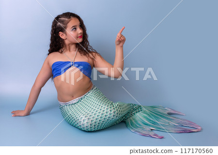 A child girl in a mermaid costume sits on a blue background 117170560