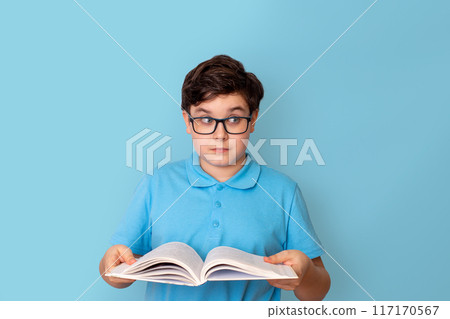 A surprised schoolboy in glasses, holding an open book in his hands, looking away 117170567