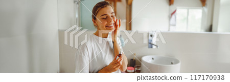 Positive female applying serum on face moisturizing and caring for skin standing in bathroom 117170938