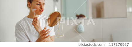 Pretty woman massaging face with jade roller and looking at round cosmetic mirror in bathroom Pretty woman massaging face with jade roller and looking at round cosmetic mirror in bathroom 117170957