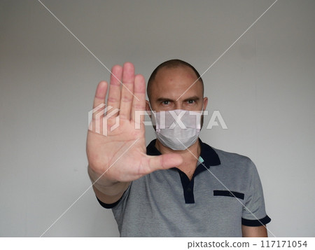 Stern looking man in medical mask shows open palm stop gesture on light background with copy space 117171054
