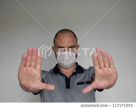 Stern looking man in medical mask shows open palm stop gesture on light background with copy space Stern looking man in medical mask shows open palm stop gesture on light background with copy space 117171055