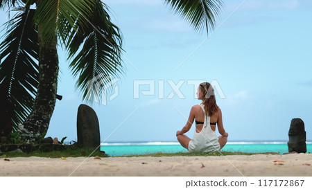 Woman sitting in yoga position on sandy beach, with her legs crossed and hands resting on her knees. The blue ocean and clear sky provide a serene backdrop as she practices mindfulness and relaxation. Woman sitting in yoga position on sandy beach, with her legs crossed and hands resting on her knees. The blue ocean and clear sky provide a serene backdrop as she practices mindfulness and relaxation. 117172867