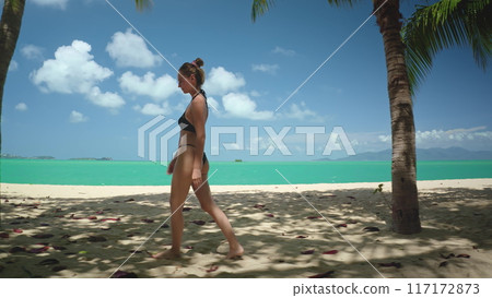 Young woman in black bikini relax walking white sand beach in Thailand. The turquoise sea water in background, blue sky and palm trees. Girl enjoy outdoor lifestyle travel on summer holiday vacation. Young woman in black bikini relax walking white sand beach in Thailand. The turquoise sea water in background, blue sky and palm trees. Girl enjoy outdoor lifestyle travel on summer holiday vacation. 117172873