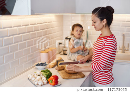 mother and cute little child daughter preparing breakfast in modern white kitchen. mom cutting bread on table. mother and cute little child daughter preparing breakfast in modern white kitchen. mom cutting bread on table. 117173055