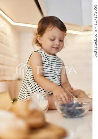 Little baby girl eating fresh blueberries from glass bowl in white kitchen 117173059
