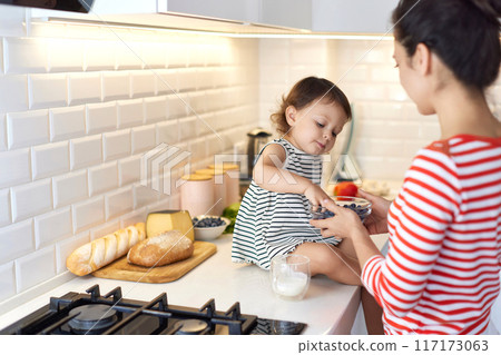 Little baby girl with her mother eating fresh blueberries in white kitchen Little baby girl with her mother eating fresh blueberries in white kitchen 117173063