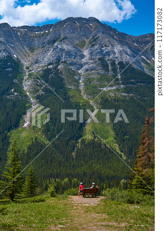 A couple sits on a bench and enjoys the view of a majestic rocky mountain in the Alberta province of Canada. 117173082