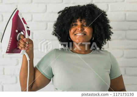 Young satisfied african american housekeeper woman wear mint t-shirt hold in hands on iron on brick wall background. Housework housekeeping concept Young satisfied african american housekeeper woman wear mint t-shirt hold in hands on iron on brick wall background. Housework housekeeping concept 117173130