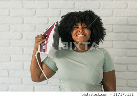 Young satisfied african american housekeeper woman wear mint t-shirt hold in hands on iron on brick wall background. Housework housekeeping concept 117173131