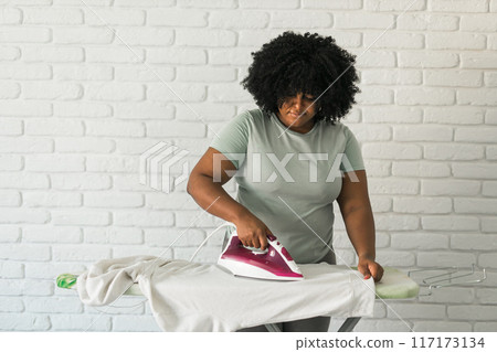 Young happy african american woman ironing clothes on ironing board at home. Copy space Young happy african american woman ironing clothes on ironing board at home. Copy space 117173134