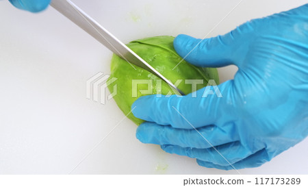 Ripe avocado cutting close up on white kitchen board. Chef hands in blue gloves mince fresh vegetable with big iron knife. Cooking, preparing ingredients in professional restaurant. Top down view 117173289