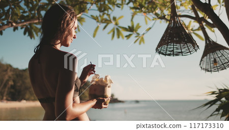 Woman enjoy tropical coconut cocktail on sunset beach. Female in bikini relax under tree in soft light sea coast landscape. Female tourist on summer vacation journey. Lifestyle holiday concept 117173310