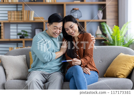 Asian couple celebrating positive pregnancy test result at home. Happy future parents enjoying moment of joy and anticipation while sitting on sofa. Expecting new baby and being joyful. Asian couple celebrating positive pregnancy test result at home. Happy future parents enjoying moment of joy and anticipation while sitting on sofa. Expecting new baby and being joyful. 117173381