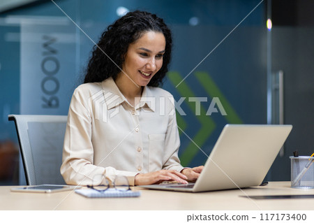 Confident businesswoman working on laptop in modern office environment. Professional female employee focusing on tasks, smiling at computer screen, displaying productivity and positivity. 117173400
