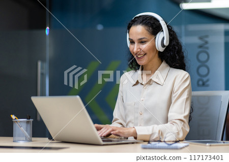 Smiling businesswoman wearing headphones working on laptop during online meeting in modern office setting. Joyful expression suggests positive communication and productivity. Smiling businesswoman wearing headphones working on laptop during online meeting in modern office setting. Joyful expression suggests positive communication and productivity. 117173401