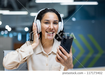 Smiling businesswoman in office environment wearing headphones, holding smartphone. Office worker engaging in conversation or enjoying music while multitasking. Smiling businesswoman in office environment wearing headphones, holding smartphone. Office worker engaging in conversation or enjoying music while multitasking. 117173402