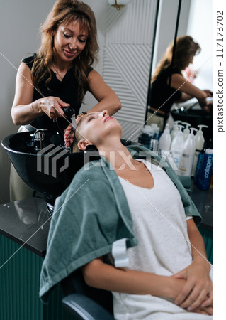 Vertical portrait of female hairdresser carefully washing hair of beautiful blonde woman in beauty salon. Beautiful customer lady lying on salon washing bed feeling relax, happy enjoy massaging head. 117173702