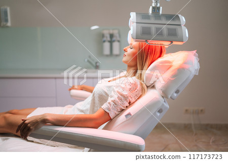 Relaxed blonde female with closed eyes lying on procedure chair having LED red light hair photodynamic therapy during hair treatment at clinic. Woman client having non-invasive type of phototherapy. Relaxed blonde female with closed eyes lying on procedure chair having LED red light hair photodynamic therapy during hair treatment at clinic. Woman client having non-invasive type of phototherapy. 117173723