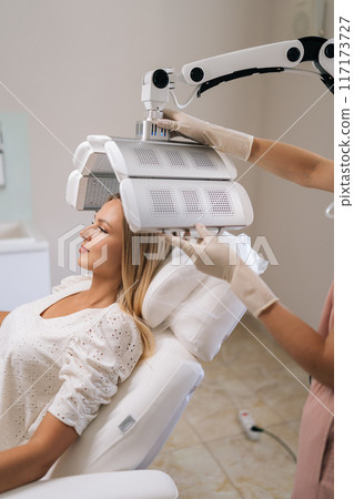 Vertical cropped shot of beautiful woman with blond hair having LED light hair PDT treatment at medical clinic. Cosmetician specialist adjusting innovation device for photodynamic therapy procedure Vertical cropped shot of beautiful woman with blond hair having LED light hair PDT treatment at medical clinic. Cosmetician specialist adjusting innovation device for photodynamic therapy procedure 117173727