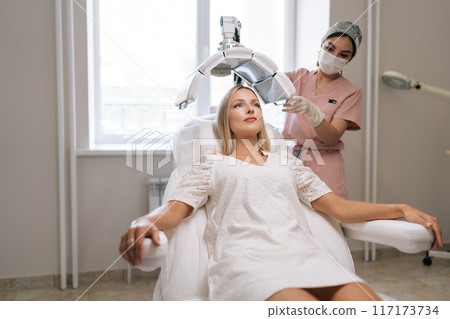 Wide shot of beautician specialist in mask connecting and adjusting innovation device for photodynamic therapy procedure during hair treatment at modern medical clinic. Concept of esthetic care. Wide shot of beautician specialist in mask connecting and adjusting innovation device for photodynamic therapy procedure during hair treatment at modern medical clinic. Concept of esthetic care. 117173734