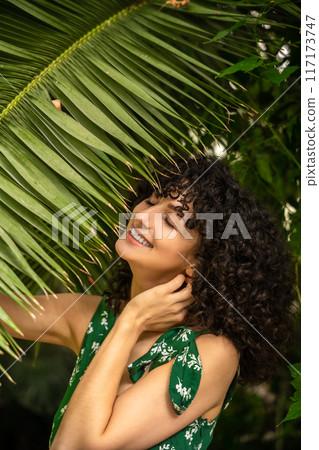 Winsome woman with curly hair and fresh greenery in the background Winsome woman with curly hair and fresh greenery in the background 117173747
