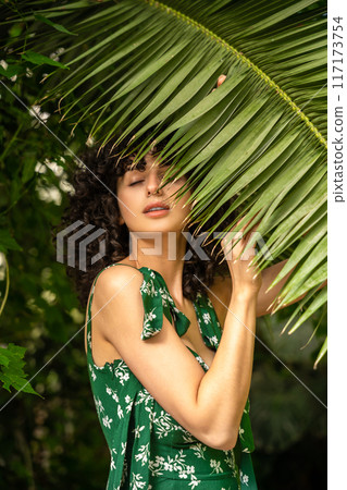 Brunette woman among tropical leaves standing with closed eyes 117173754