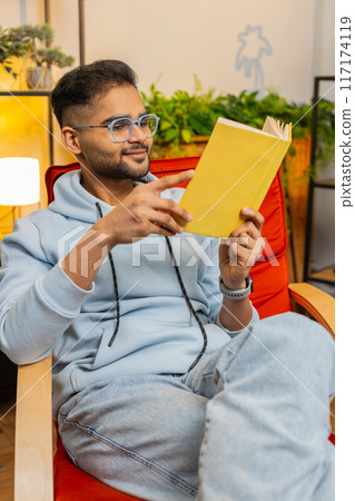 Indian man reading interesting book turning pages smiling enjoying literature taking a rest on chair Indian man reading interesting book turning pages smiling enjoying literature taking a rest on chair 117174119