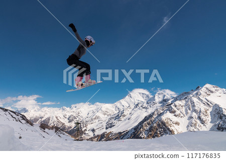 A young woman in a helmet makes a ski jump high in the mountains. Winter sport. Snowboarder woman doing a trick A young woman in a helmet makes a ski jump high in the mountains. Winter sport. Snowboarder woman doing a trick 117176835