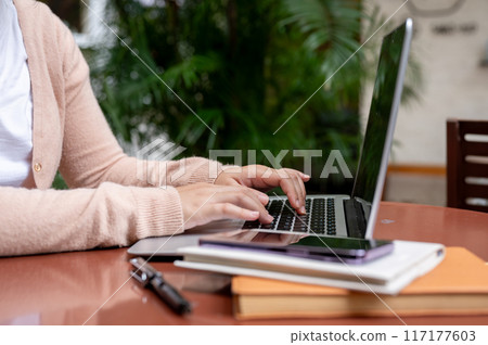 A woman working on her laptop, typing on the laptop keyboard, sitting at a table in a garden. A woman working on her laptop, typing on the laptop keyboard, sitting at a table in a garden. 117177603