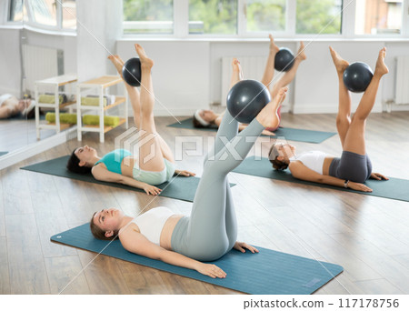 Lying on their backs, young girls hold Pilates ball between their knees and perform leg exercises Lying on their backs, young girls hold Pilates ball between their knees and perform leg exercises 117178756
