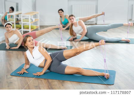 Girls perform exercises lying on side with legs straightened and narrow elastic band on shins Girls perform exercises lying on side with legs straightened and narrow elastic band on shins 117178807