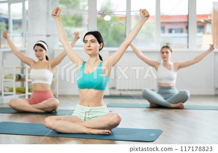 Girl conducts Pilates class for girls sitting with elastic band in hands above head makes body bends 117178823