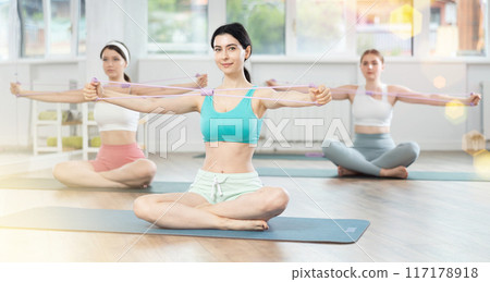 Girl conducts Pilates class for girls sitting with elastic band in hands above head makes body bends 117178918