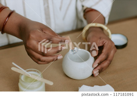 Person stirring mixture in a ceramic container while preparing skincare products at home studio. Person wearing white clothing and bracelets, emphasizing hands and motion 117181091