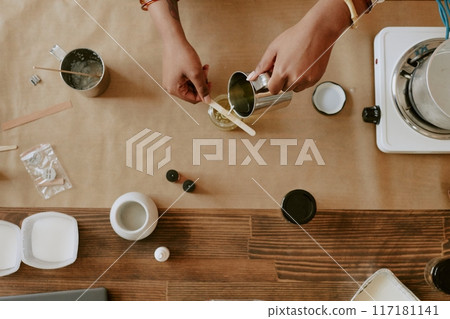 Top-down view showing hands engaged in stirring ingredients in various containers on wooden surface with crafting tools surrounding setup Top-down view showing hands engaged in stirring ingredients in various containers on wooden surface with crafting tools surrounding setup 117181141