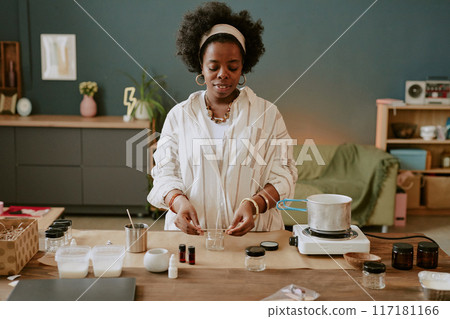 Black woman with afro hairstyle carefully mixing ingredients for handmade candles on wooden table in cozy home studio filled with various supplies and equipment 117181166